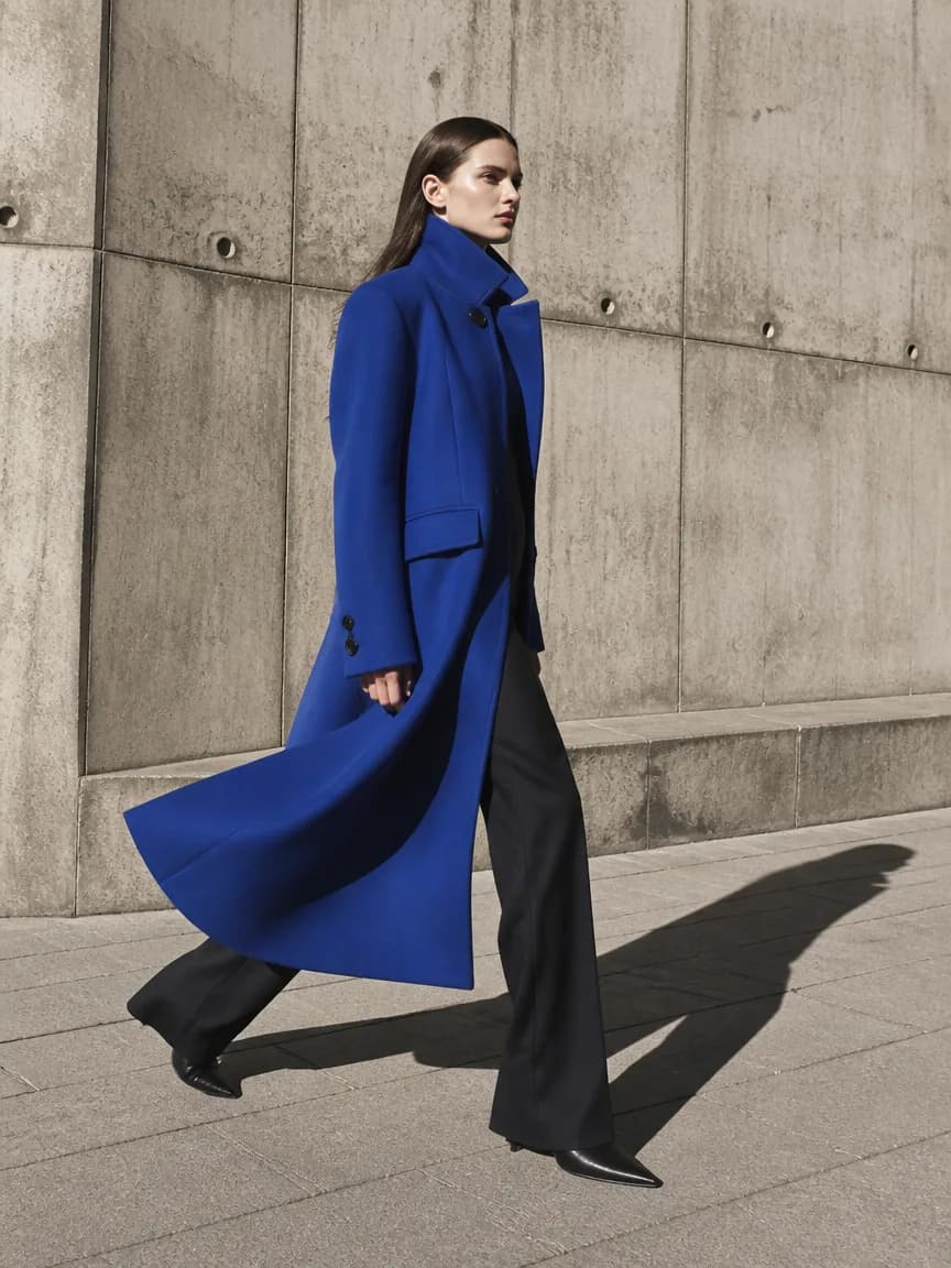 Full-length editorial of a model in a cobalt coat walking past a concrete wall
